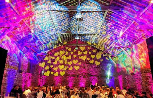 Éclairage de mariages dans une grange en pierre : projecteurs LED multicolores (rose, bleu, violet) illuminent les poutres apparentes et les murs en pierre, avec une projection de cœurs dorés en fond de salle.